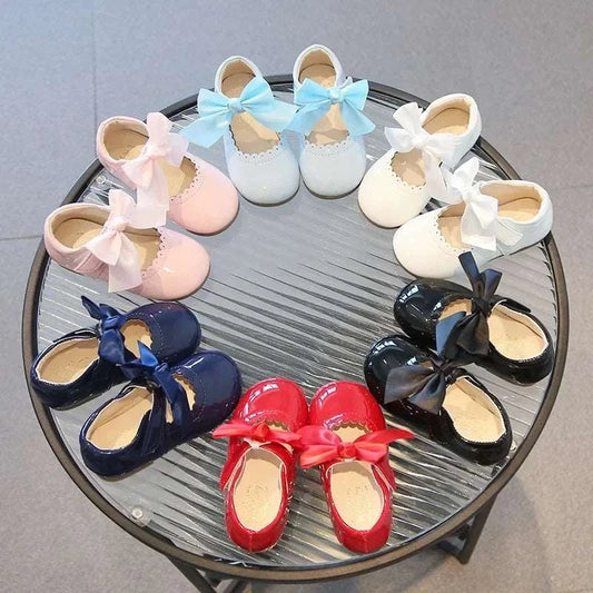 Colorful little toddler girl leather dress shoes with bows in pink, blue, white, navy, red, and black arranged in a circle on a glass table